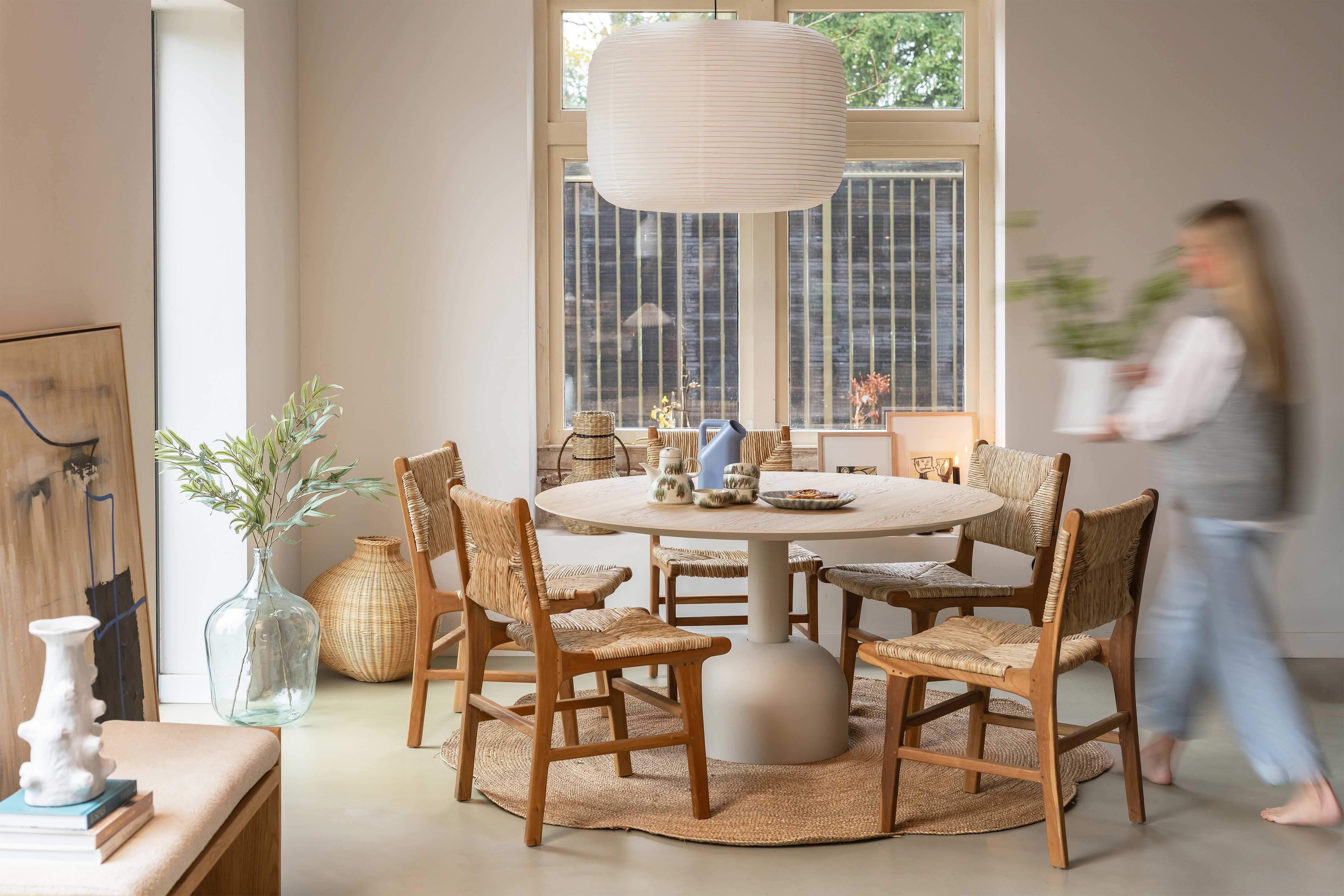 Ronde eettafel met houten rieten stoelen eromheen en blonde vrouw die vaas met bloemen neerzet