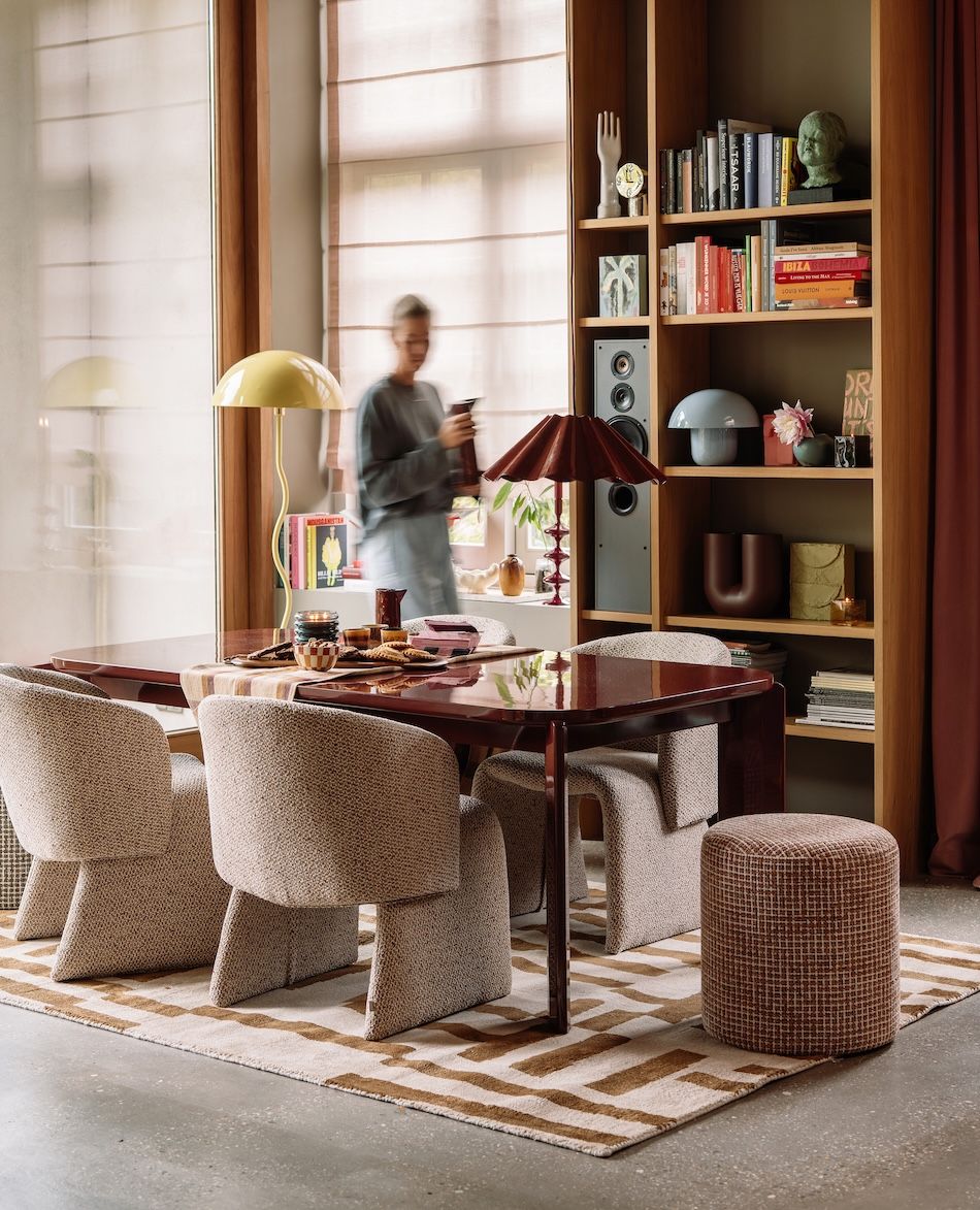 tafel met eetkamerstoelen, poefje en vloerkleed. Vrouw loopt door ruimte
