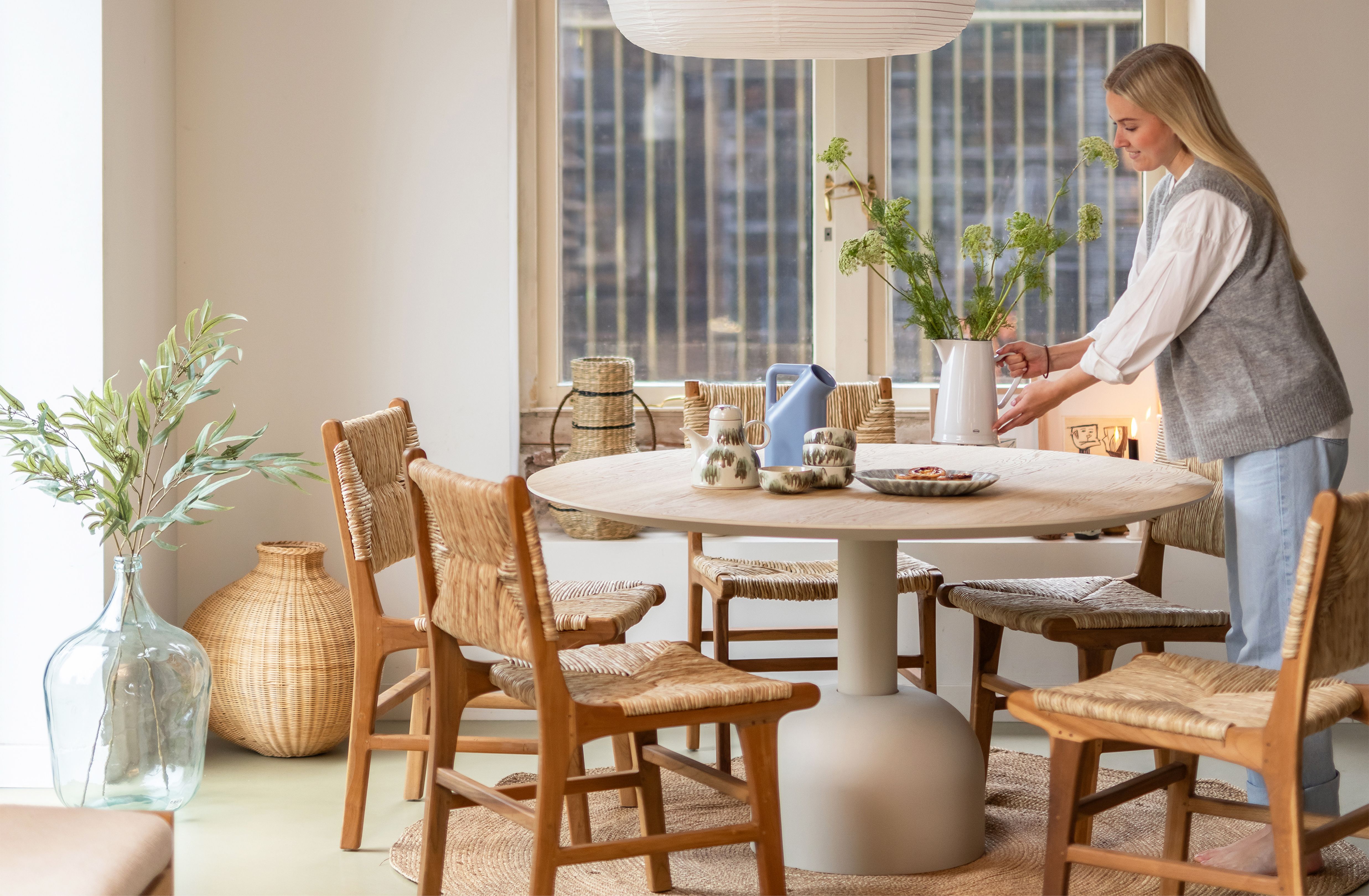 Ronde eettafel met houten rieten stoelen eromheen en blonde vrouw die vaas met bloemen neerzet